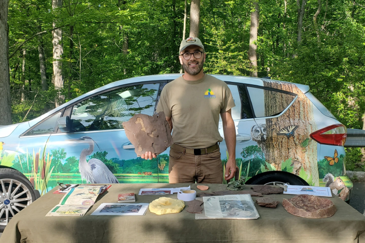 dinosaur fossil held up by a man with dinosaur items on a table with dinosaur painted car in the background
